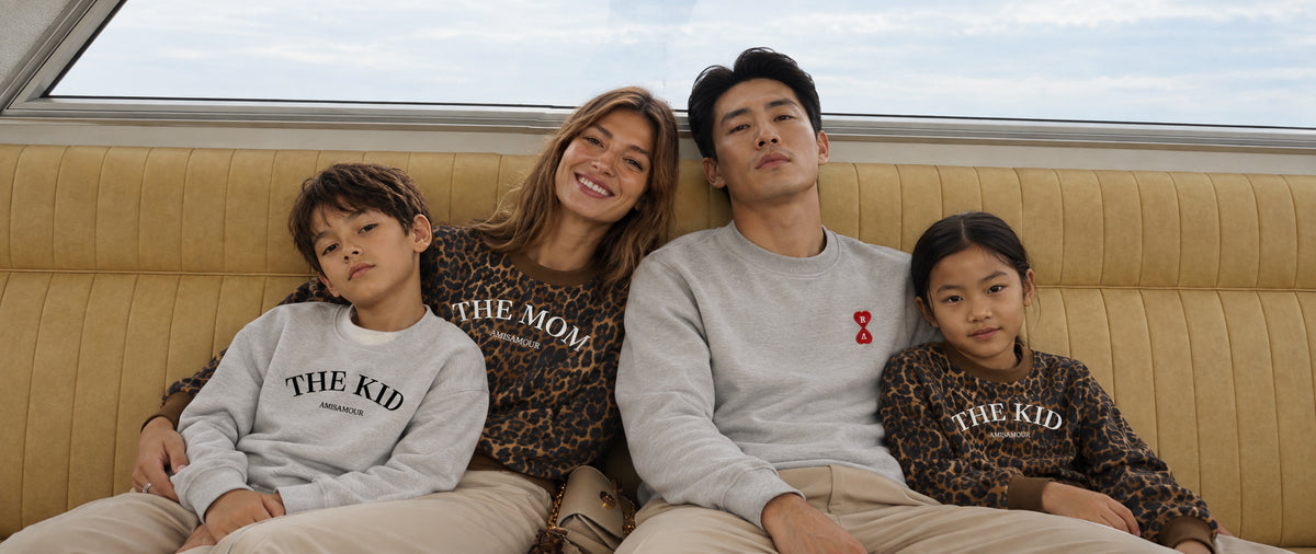 A family of four wearing a matching 'The Mom & The Kid' apparel collection, sitting together on a retro-style beige leather bench. The set includes a man and a woman in light gray sweatshirts, and two children in leopard-print sweatshirts and sweatpants, all featuring 'THE MOM & THE KID' text on the front.