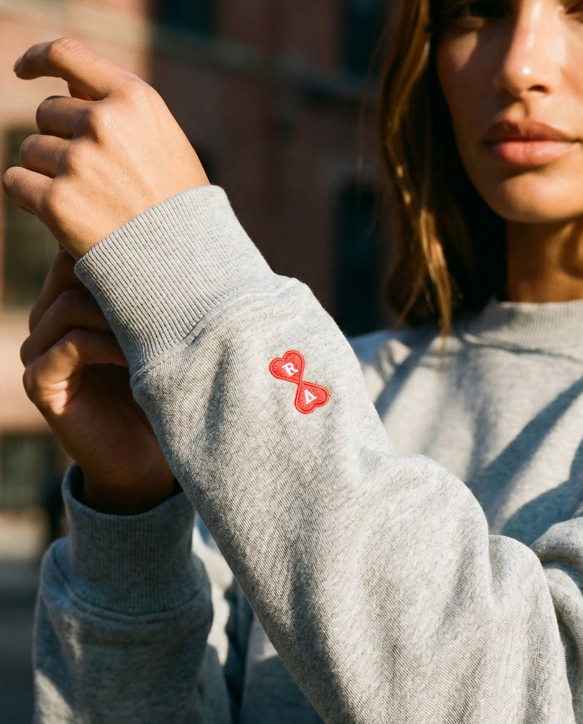 Person wearing a grey AmisAmour sweatshirt with a small red infinity heart logo embroidered on the sleeve.